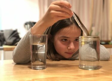 Ein Mädchen beobachet genau einen naturwissenschaftlichen Versuch, das es mit Wasser, einer Pipette und zwei Glasgefäßen selbst durchführt.